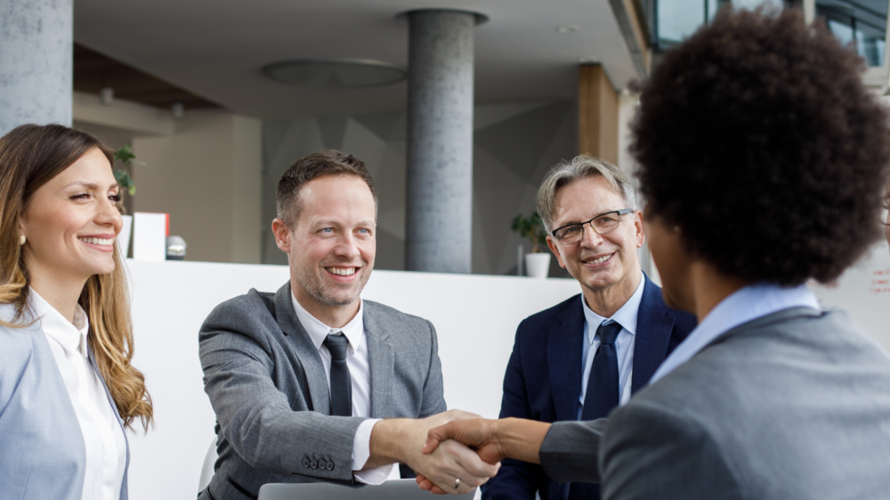 Group of professionals in formal attire seated in a modern office setting, with one person extending a handshake to another, symbolizing talent acquisition, recruitment, and collaborative hiring processes.