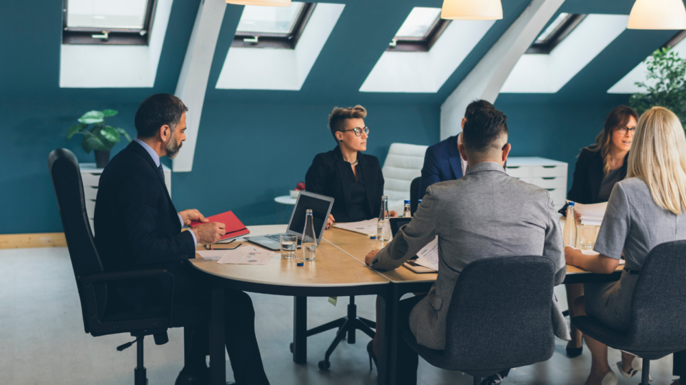 A modern conference room with a round wooden table where several professionals are seated, engaged in discussion. The setting features slanted ceiling beams, large skylight windows allowing natural light, and pendant lights hanging above the table