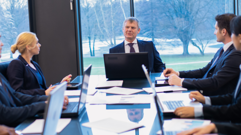 A formal meeting in a modern conference room with large glass windows overlooking trees and natural scenery. Several professionals in business attire are seated around a polished table with laptops and documents, engaged in discussion. The setting represents environmental law and corporate decision-making related to regulatory compliance and sustainability.