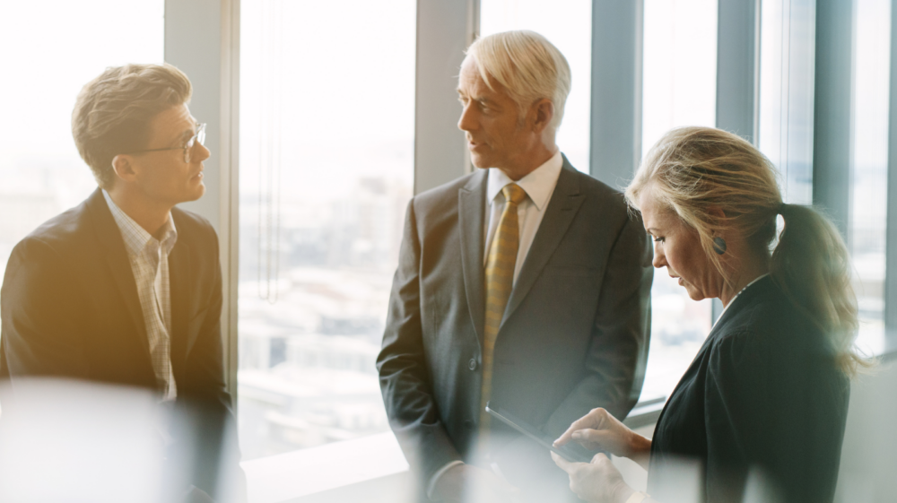 Three professionals standing near large floor-to-ceiling windows in a modern office, engaged in a discussion.