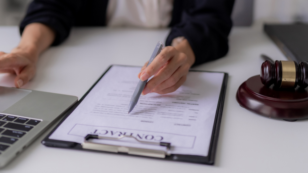A professional seated at a desk reviewing a contract document on a clipboard, with a pen in hand and a laptop nearby. A judge’s gavel rests on the desk, symbolizing legal authority and intellectual property rights. The setting conveys a formal office environment focused on legal documentation and compliance.