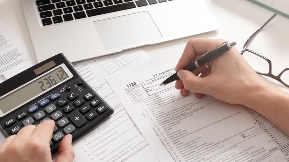 A workspace with tax forms, a laptop displaying financial documents, a calculator showing numbers, and a person’s hands writing on a form while performing calculations. Nearby are binders and paperwork, representing tax preparation and financial documentation.