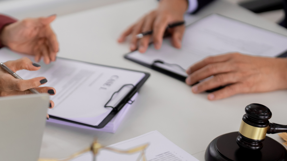 A close-up view of a legal meeting table with documents labeled “CASE” on clipboards, a judge’s gavel, and scales of justice. Two individuals are engaged in discussion, reviewing paperwork and using a laptop, representing bankruptcy restructuring and legal proceedings.