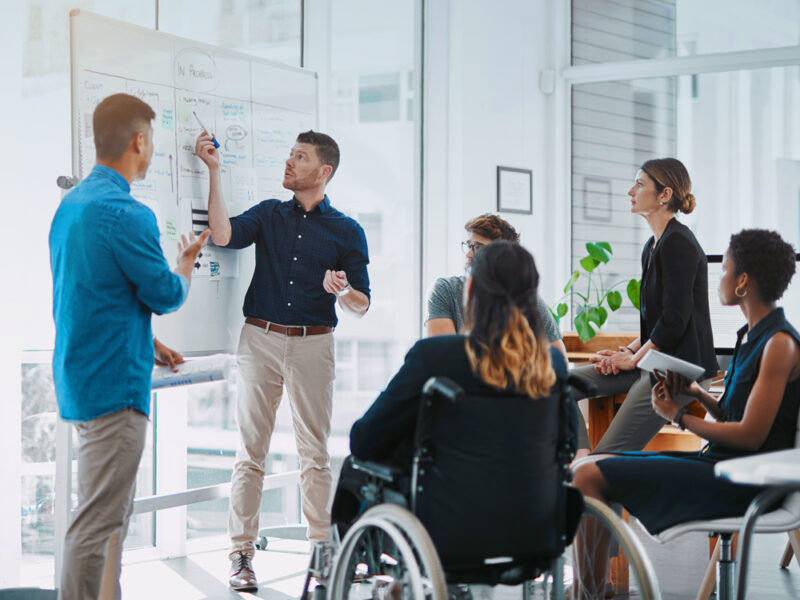 Team of professionals, including a person in a wheelchair, collaborating around a whiteboard during a financial planning session