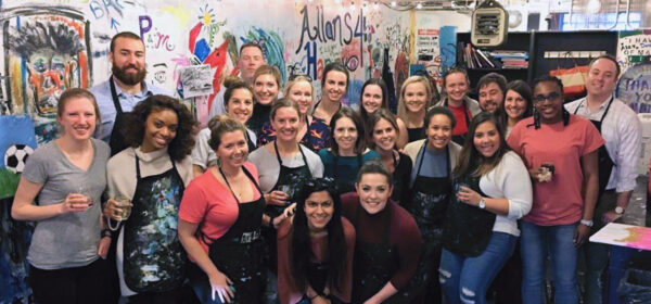 Group of recruiters enjoying a paint night team-building event, posing in front of colorful murals while wearing aprons and smiling in a collaborative, creative atmosphere