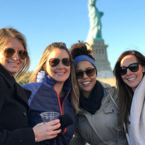Group of recruiters smiling in front of the Statue of Liberty during a team outing in New York City, showcasing camaraderie and team culture in the staffing industry