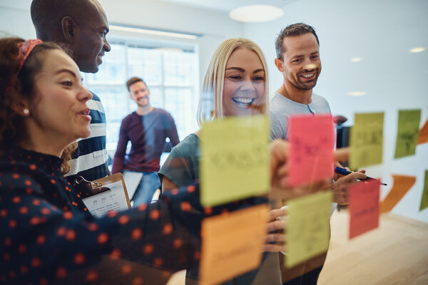 A team of professionals smiling and collaborating in a modern office, organizing colorful sticky notes on a glass wall during a strategy session.