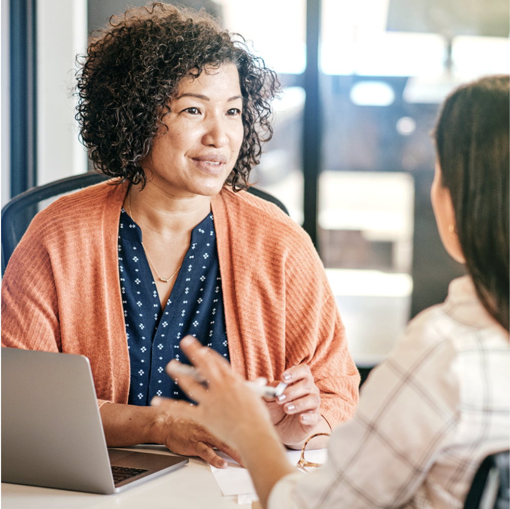 A middle aged mixed-race woman in a peach blazer with curly dark hair interviewing with a white woman wearing a light plaid shirt, who has straight dark hair