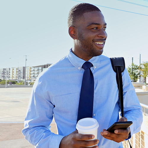 A mid-thirties African-American man outside, wearing a light blue shirt and a dark blue tie, holding a cup of takeout coffee and a phone